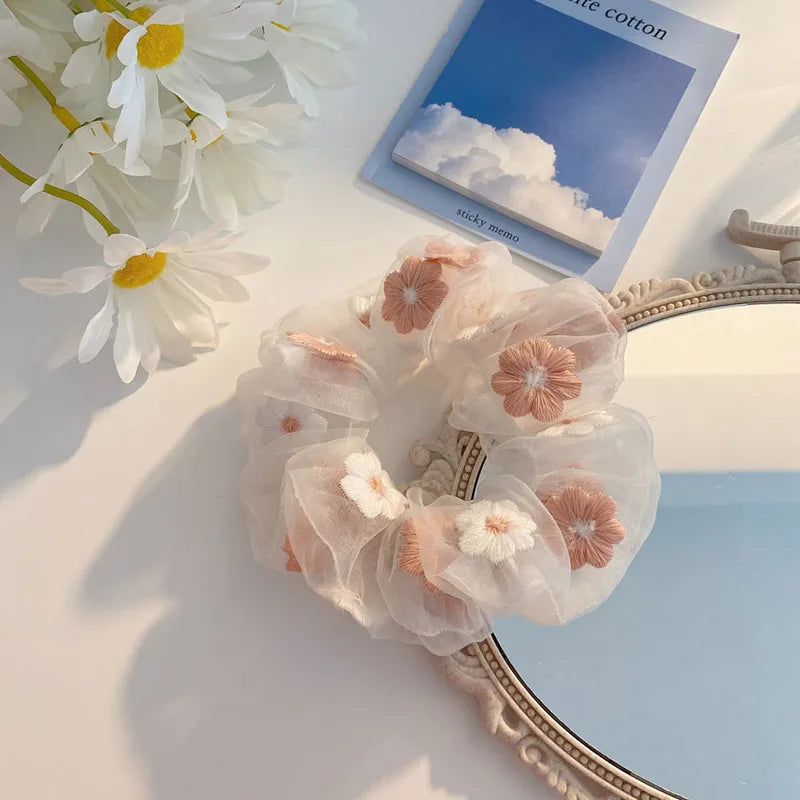 Decorative hair scrunchies with floral design on a light surface with flowers and a card in the background.