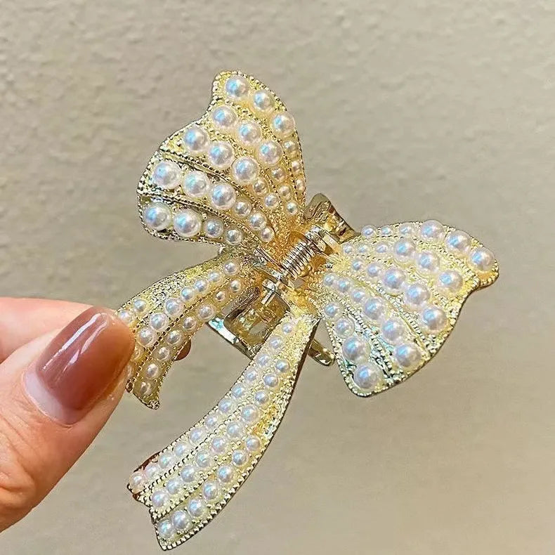 Gold bow-shaped hair clip with pearl details held by a hand against a neutral background