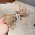 Gold bow hair clip with decorative elements held by a hand on a neutral background